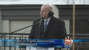 John Munchmeyer wearing coat, tie, earmuffs; standing outdoors at lectern with microphone, addressing gathering; TV screen capture; text marquee reads: 'Bill of Rights Anniversay' and 'Charlottesville' and station ID 'NBC 29'