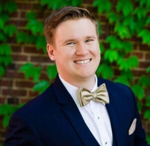 Bennett Morris grinning broadly wearing dark blue suit with beige bow tie, in front of ivy-covered brick wall (color photo)