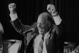 Mark Tippetts standing at microphone, speaking and smiling, fists raised, wearing suit and tie (black & white photo)