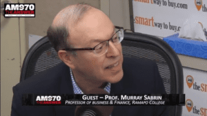 Murray Sabrin seated in radio studio, speaking on camera, microphone in front of him, wearing eyeglasses and dark sportcoat, turned to off-camera radio host at his left, text on screen identifying Sabrin as 'Guest -- Prof. Murray Sabrin' and 'Professor of Business & Finance, Ramapo College', also station ID of "AM970 THE ANSWER' (color image)