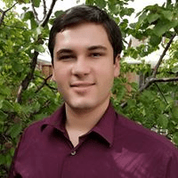 Brandon Nelson portrait, wearing burgundy shirt with open collar, clean-shaven, dark hair, standing outdoors in front of plants (color photo)