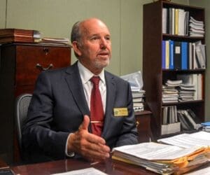 Jeff Hewitt seated behind desk in office with dark wooden furniture, speaking to someone standing off camera, gesturing, wearing suit, tie, and his ‘City of Calimesa mayor’ name badge (color photo)
