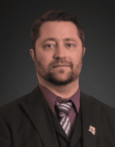 Jared Lord head shot portrait, wearing dark 3-pc suit with lapel pin, aubergine shirt, striped tie, sporting beard & mustache, expression of repose, plain dark grey backdrop (color photo)