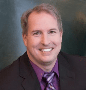 Josh Welter head shot portrait, wearing dark suit, aubergine shirt, striped tie, smiling at camera, bluish-green backdrop (color photo)