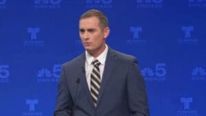 TV screen image of Kash Jackson speaking on stage at a mic wearing grey suit white shirt striped tie, blue backdrop featuring logos of Telemundo Chicago and NBC (peacock) Chicago Channel 5 (color image)