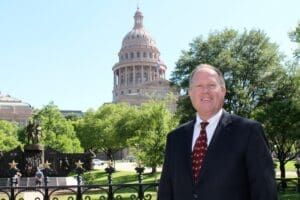 Mark Tippetts standing on verdant front grounds of Texas capitol building on sunny day, wearing dark suit white shirt maroon tie smiling at viewer (color photo)